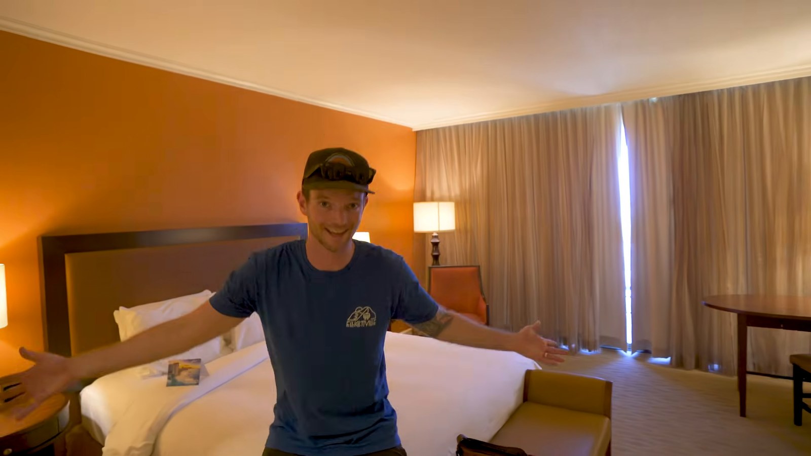 A man spreads his arms in a hotel room with a king bed, a desk and drawn curtains.