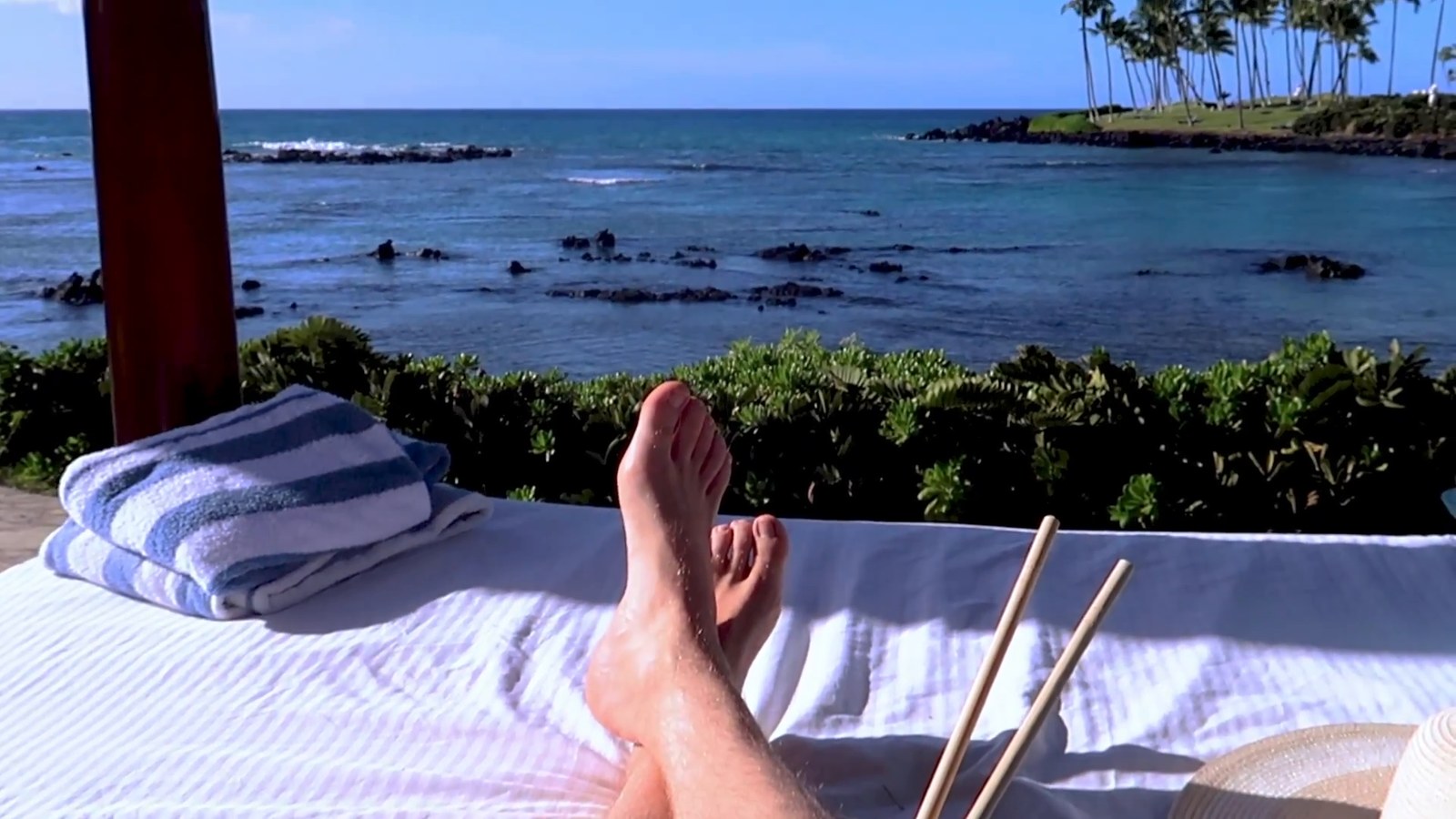 A white day bed in a shaded cabana looking out over the ocean and lava rocks, with someone's feet in the foreground.
