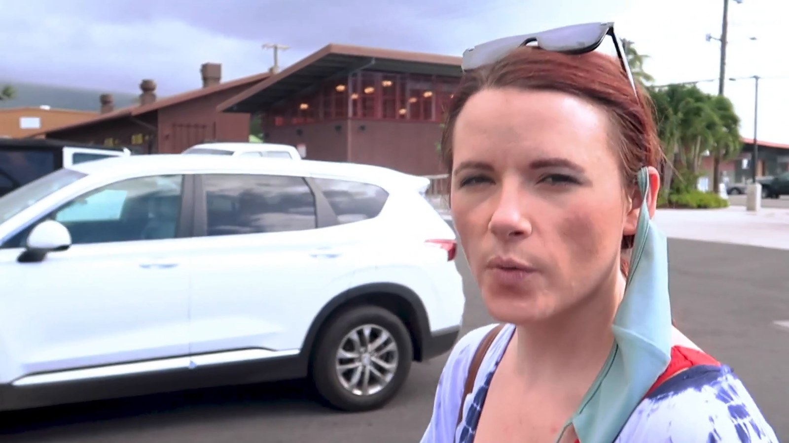 A woman with a face mask pulled down standing in a Kona parking lot beside a white SUV, with a low wooden building behind her.
