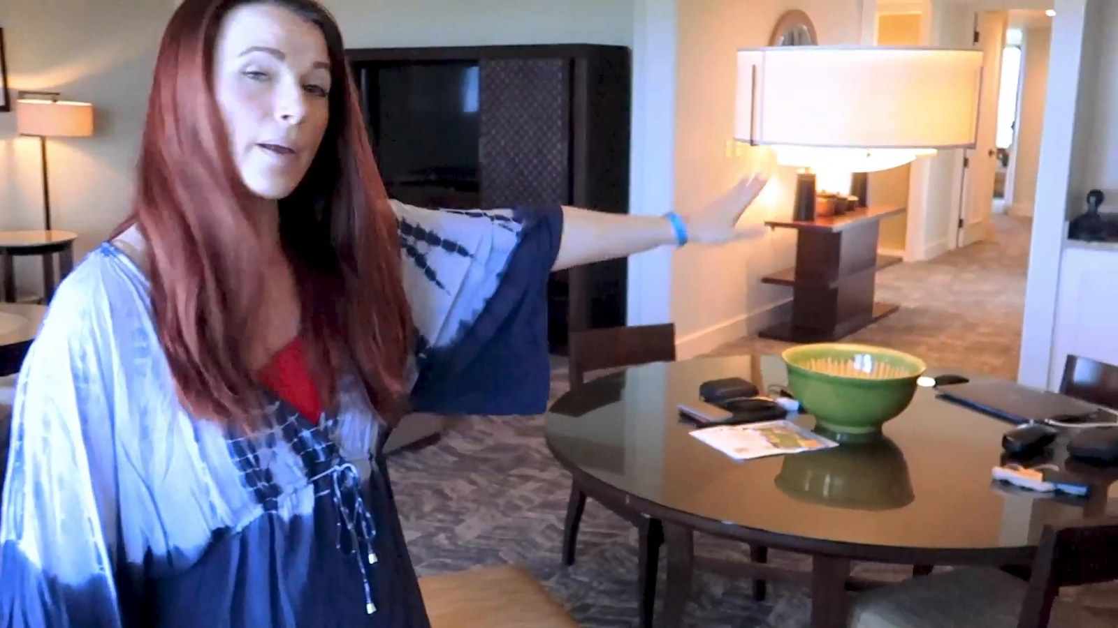 A woman gesturing across the living area of the suite, with a round dining table, a bowl, lamps and a TV behind.