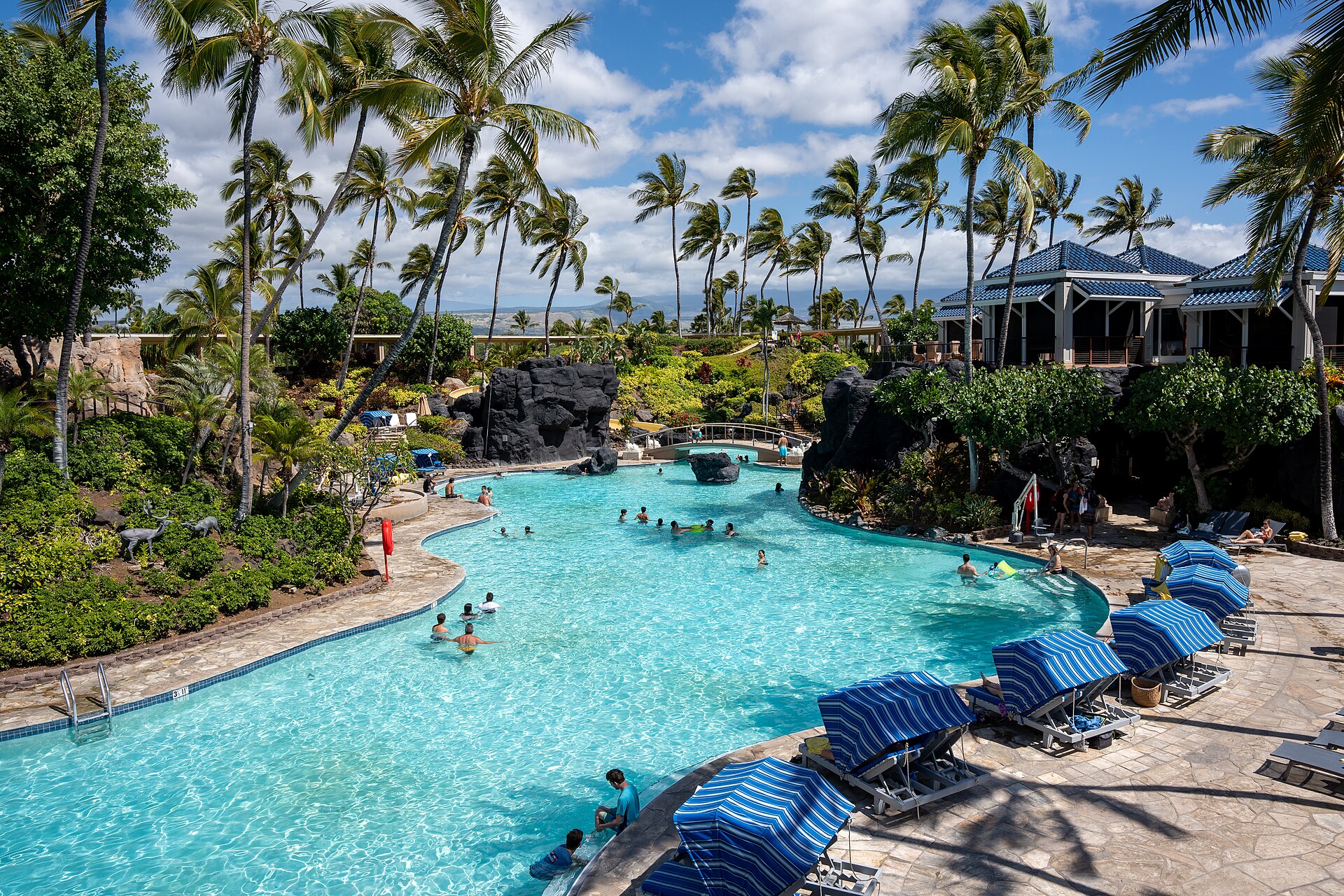 A winding resort pool with blue umbrellas, rock features, a footbridge and palm trees at Hilton Waikoloa Village.
