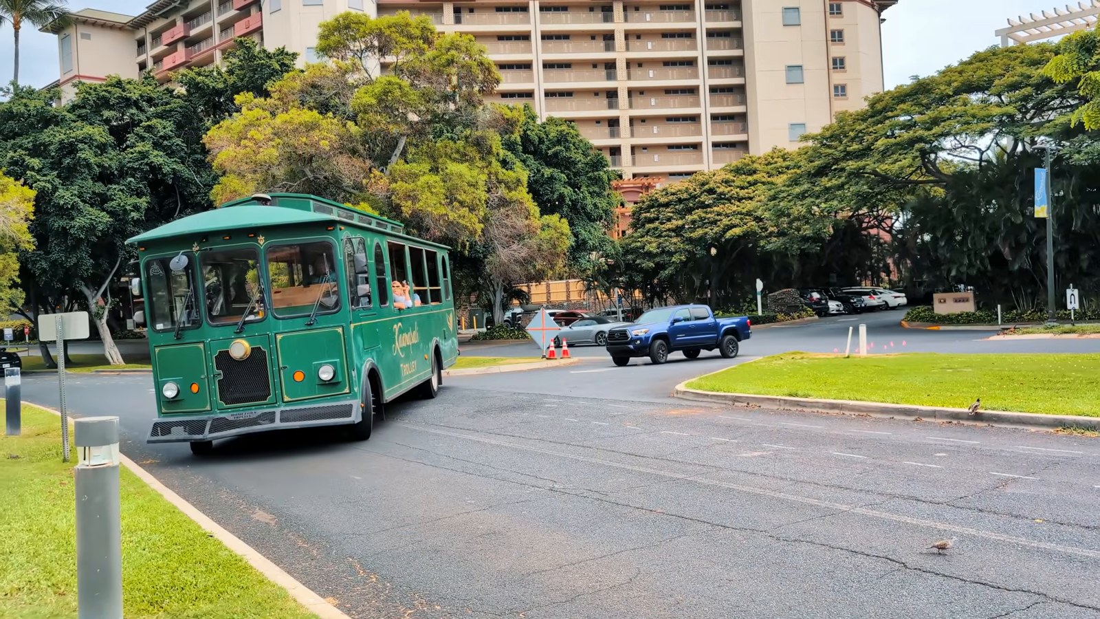 A green trolley-style bus turning on a tree-lined road in the Ka'anapali resort area with a hotel tower behind it.