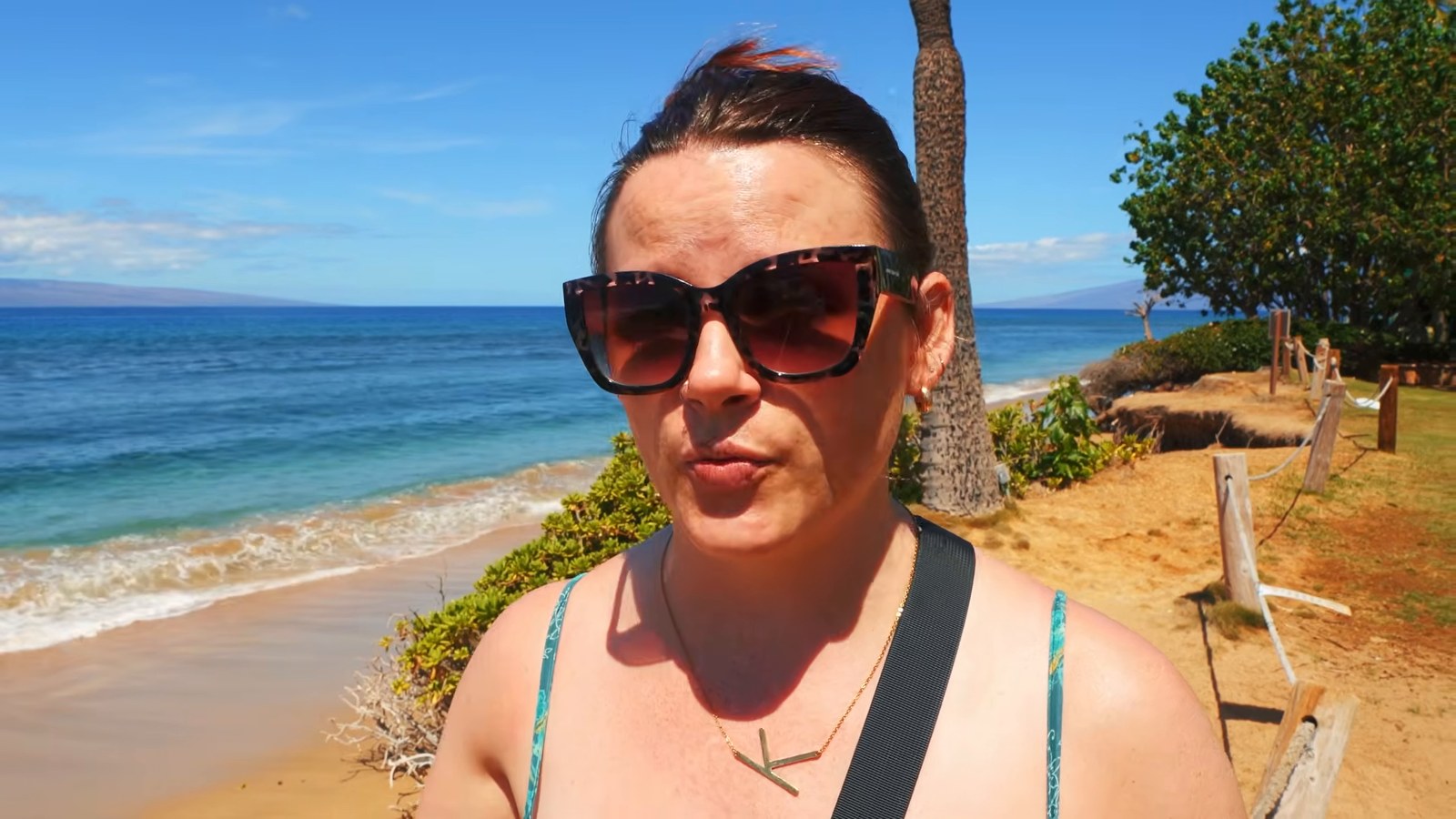A woman in sunglasses on the Ka'anapali shoreline with a rope fence at the dunes and the ocean behind her.