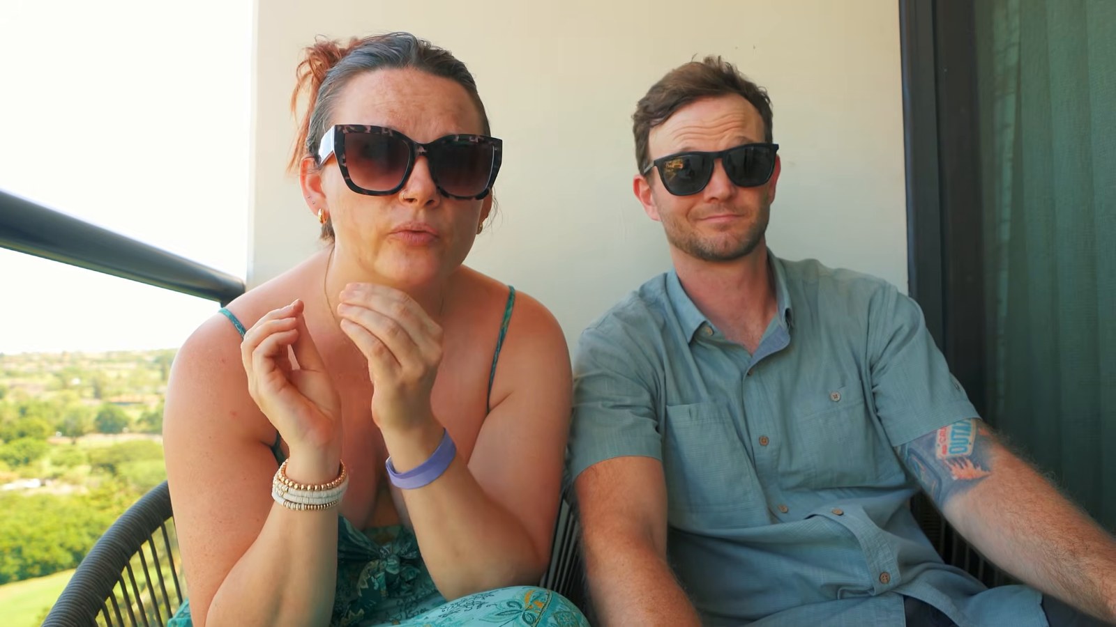 A woman and a man in sunglasses sitting together on a balcony chatting to the camera, with green grounds behind them.
