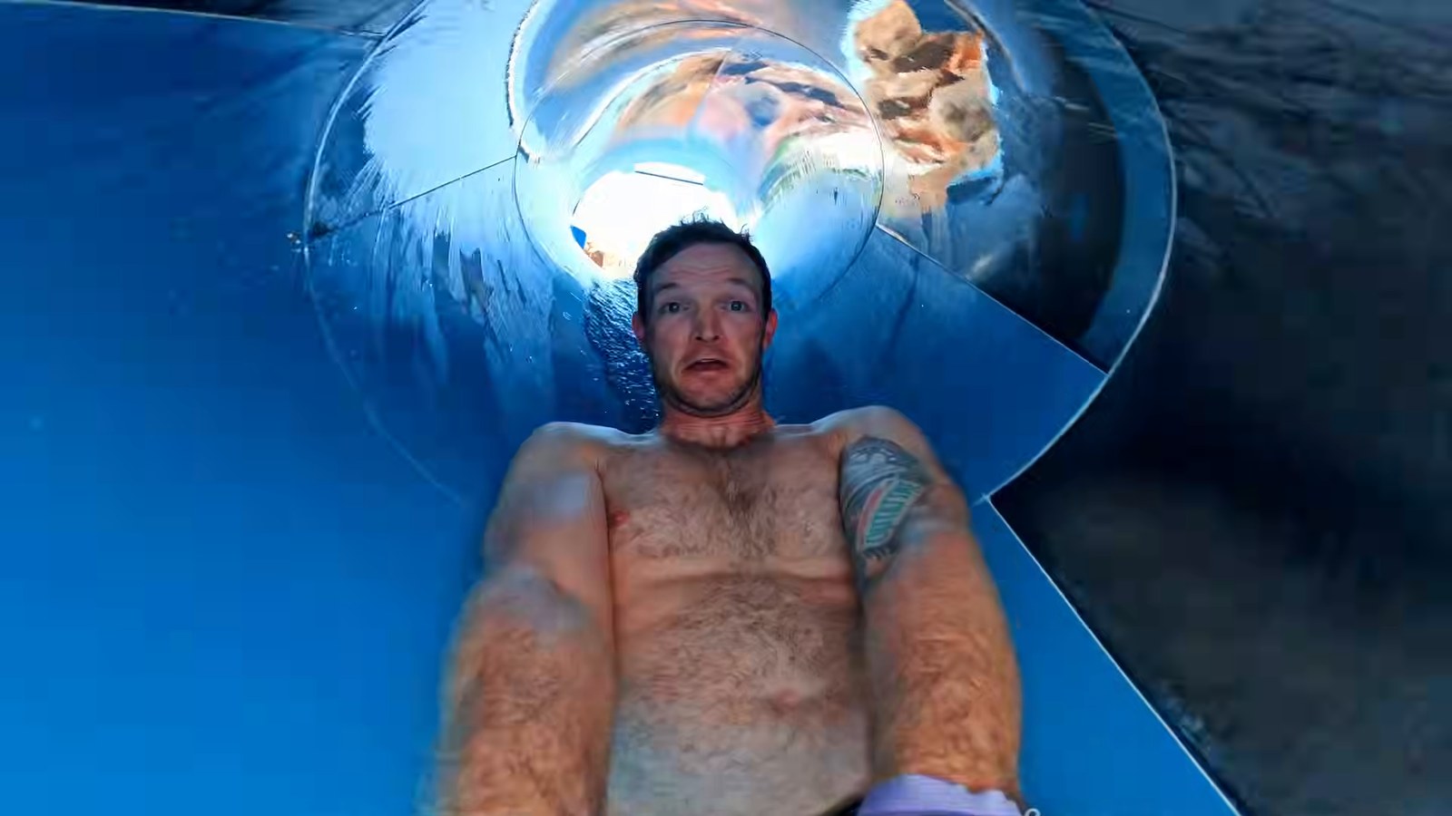 A man looking surprised as he rides down a blue lava-tube water slide, shot from a camera at his feet.