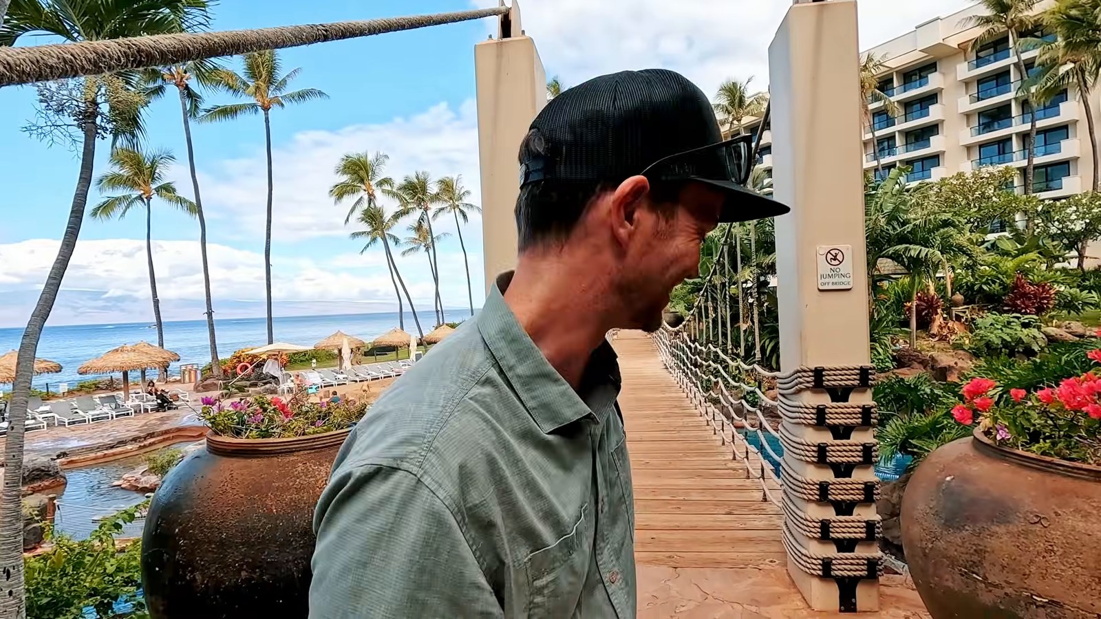 A man in a cap looking at a wooden suspension bridge over a pool, with palms, flowers and the ocean in the background.