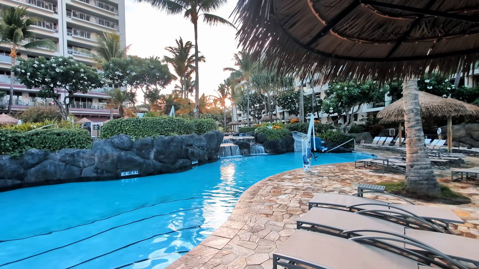 A curving pool with a rock waterfall and lounge chairs under thatched umbrellas at the Hyatt Vacation Club next to Hyatt Regency Maui.