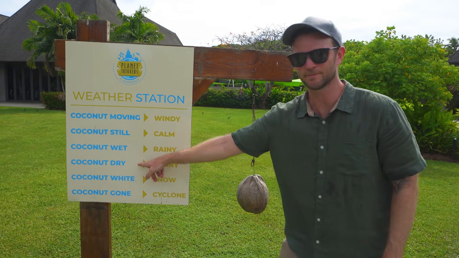 A man pointing at the resort's hand-painted 'Weather Station' sign with a hanging coconut, part of the Planet Trekkers kids' club area.