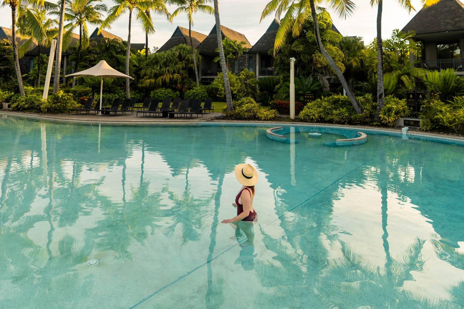 A swimmer in a straw hat wading through the resort's turquoise pool, with palm trees and thatched-roof villas behind.