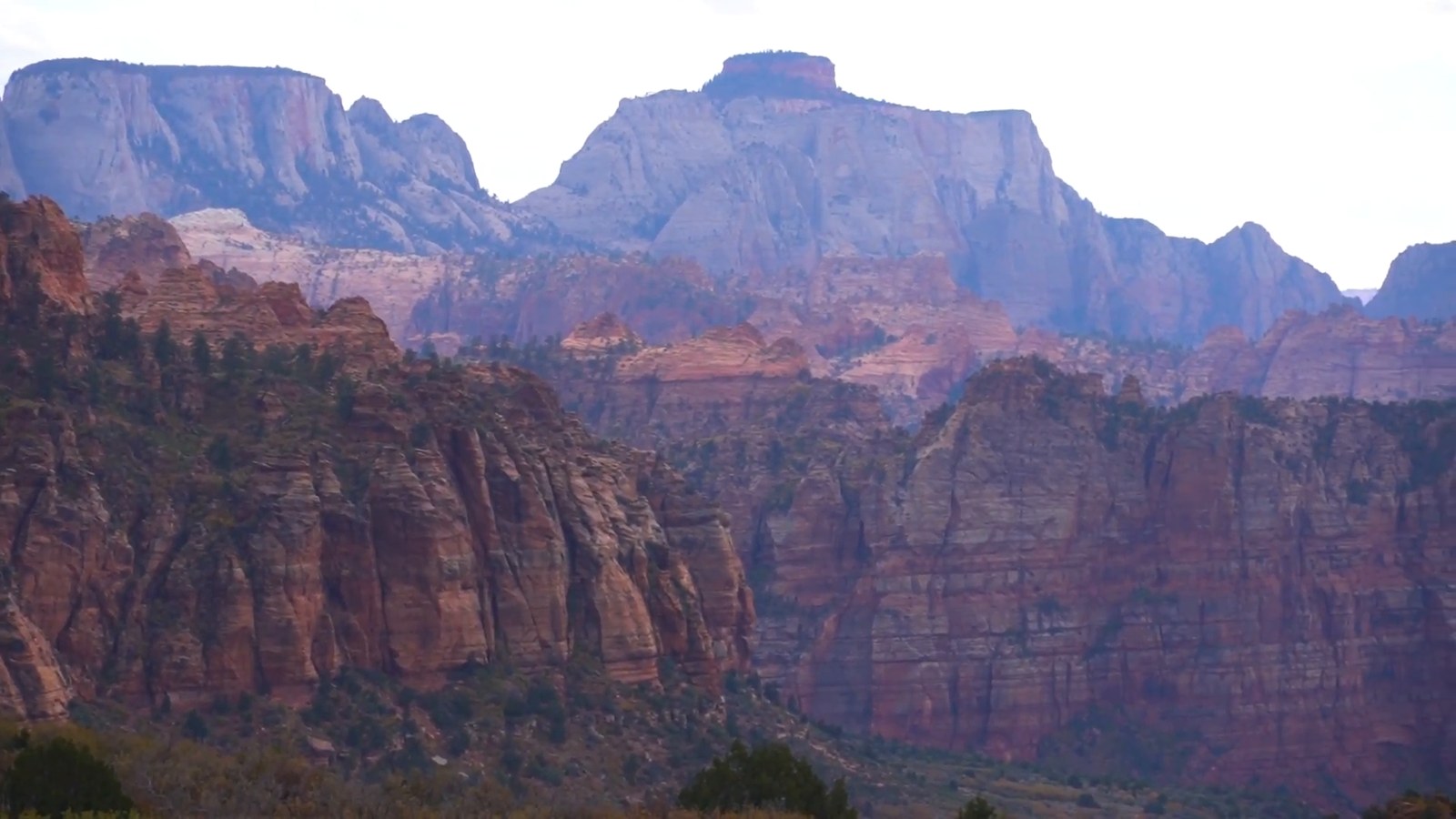 Layered red and purple sandstone cliffs and distant peaks in Zion's backcountry under a pale sky.