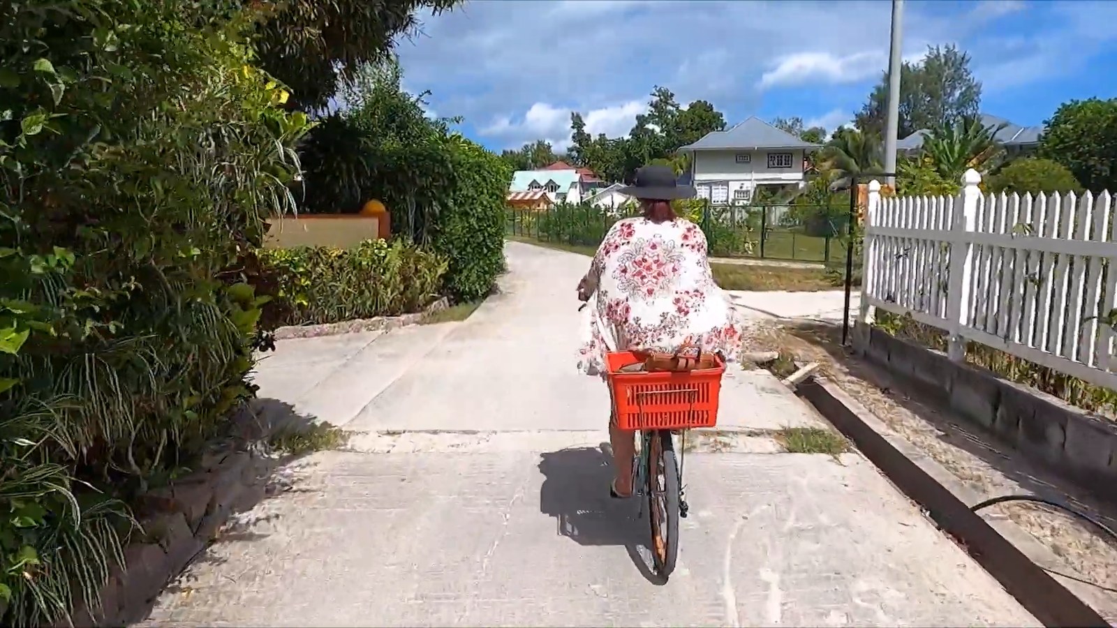 A woman in a floral top and wide-brimmed hat cycles away along a quiet concrete lane lined with hedges and small houses on La Digue.