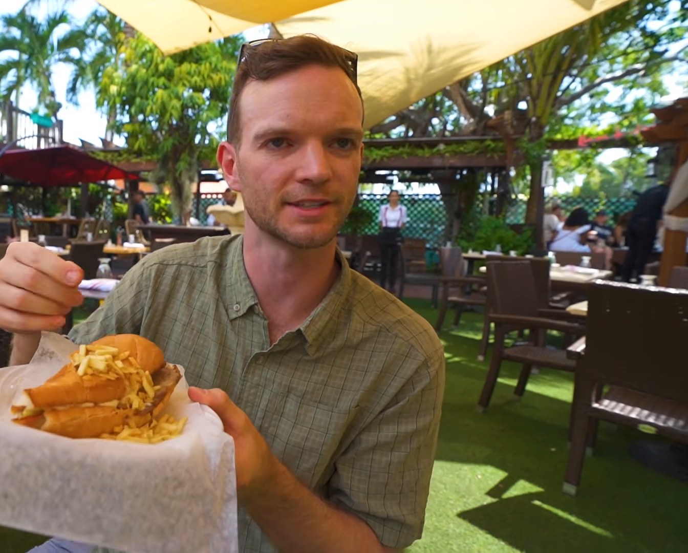 A man in a green shirt holds a Cuban sandwich with fries on a paper-lined tray in an outdoor restaurant courtyard.