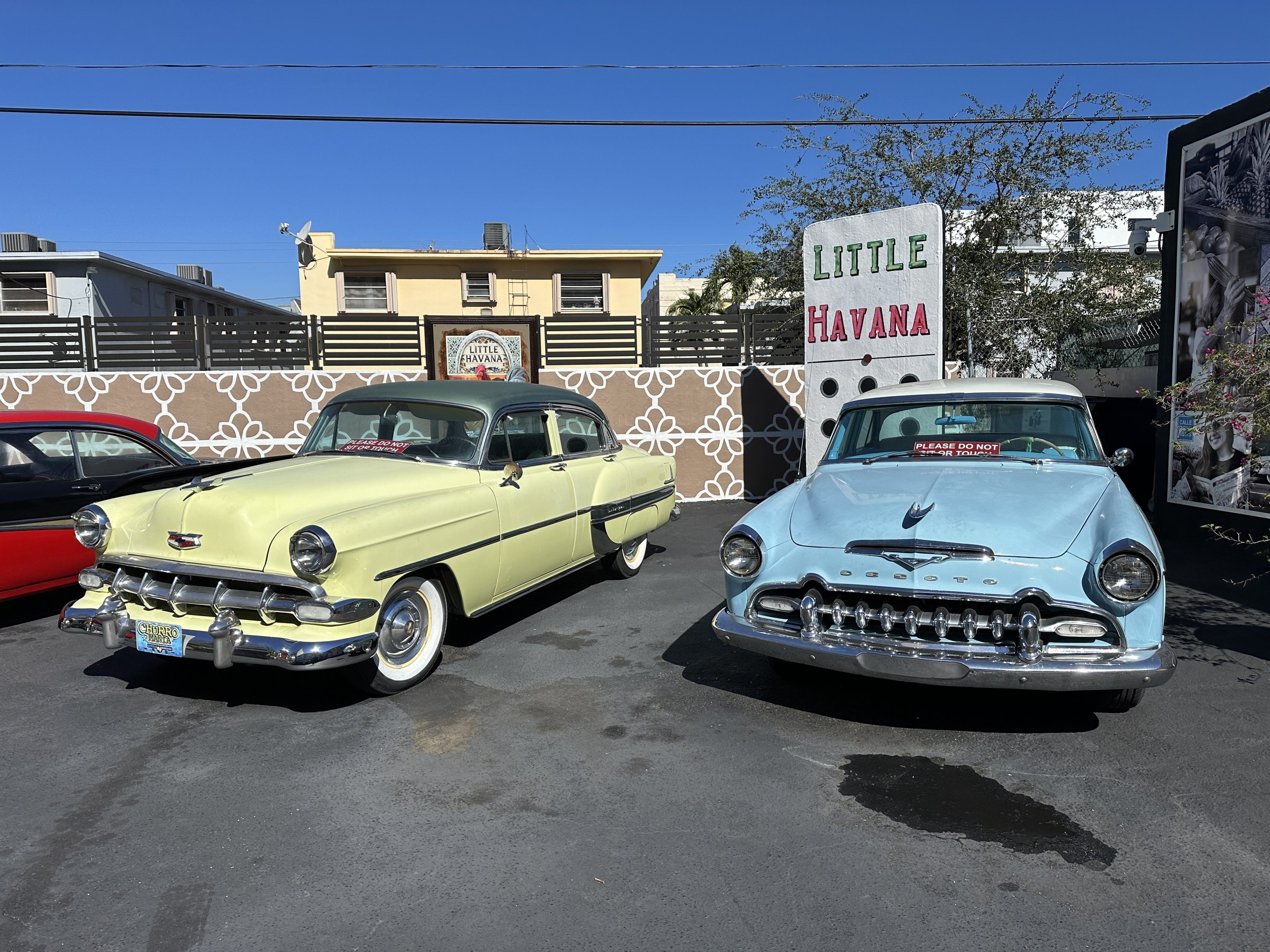Two vintage cars, one pale yellow and one light blue, parked in front of a Little Havana domino sign on Calle Ocho.