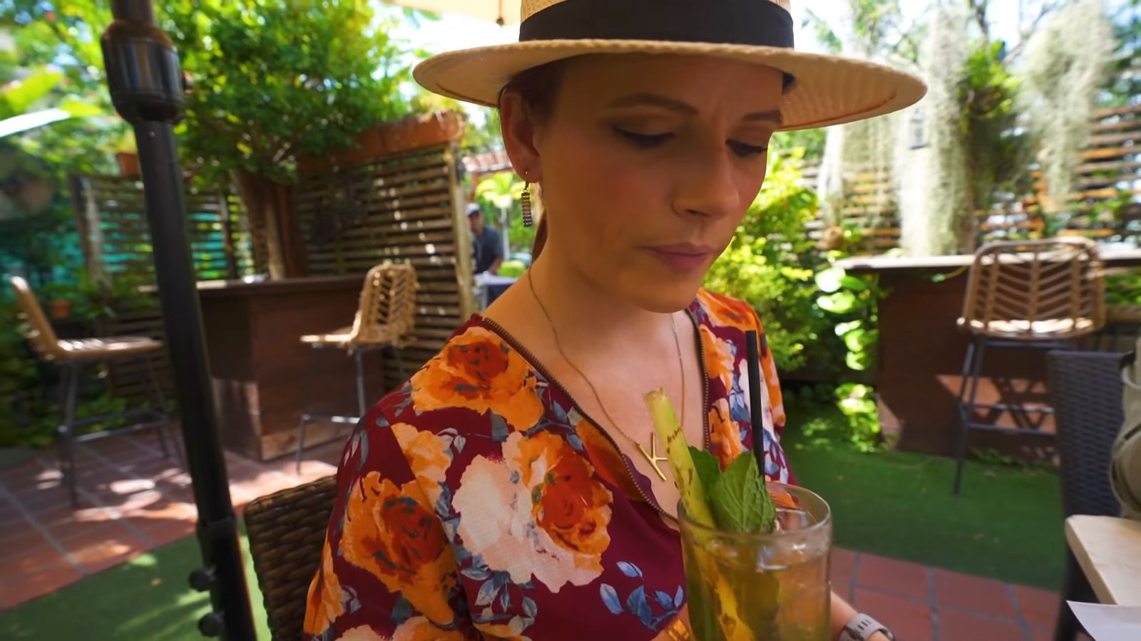 A woman in a straw hat looks down at a mojito garnished with mint in a leafy outdoor courtyard.
