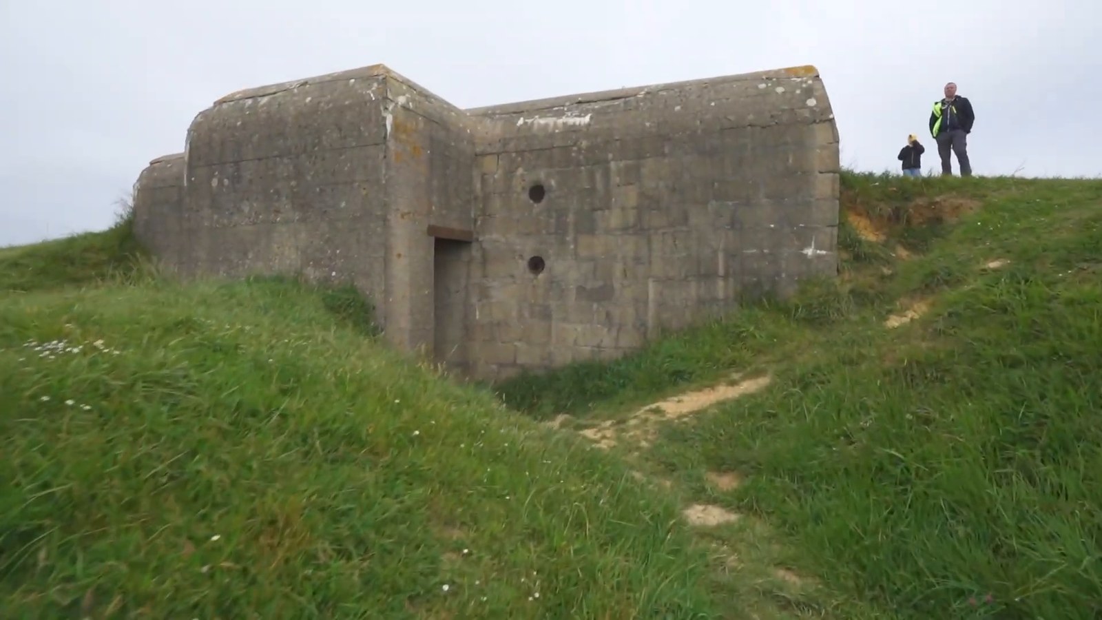 A German gun casemate of grey concrete on a grassy rise at Longues-sur-Mer under an overcast sky.