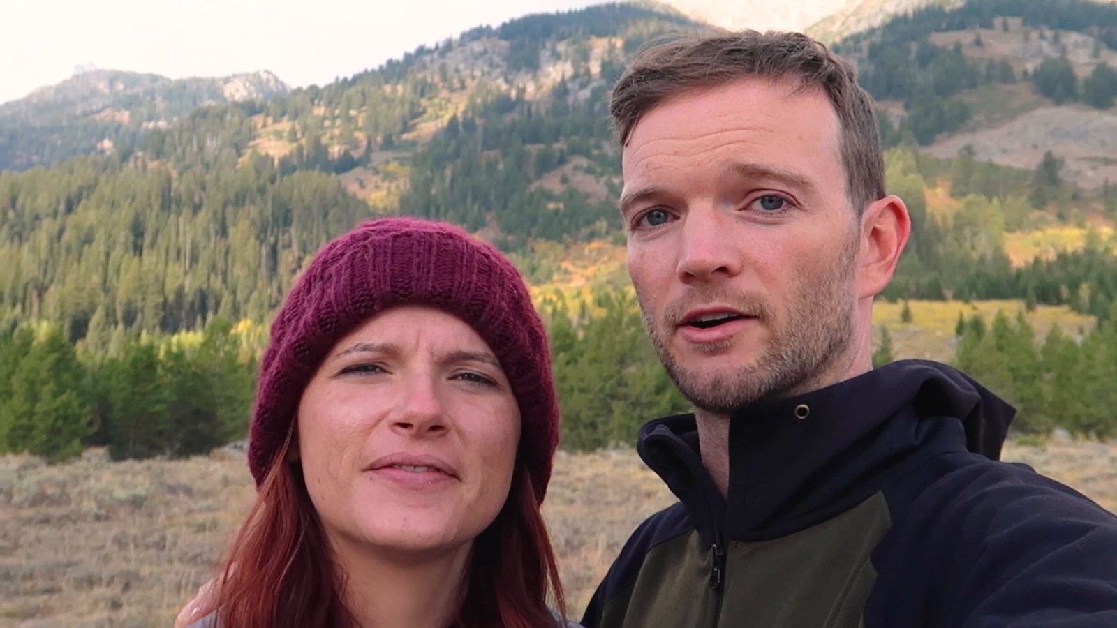 Two hikers in beanies stand in front of forested hills and mountain ridges in the early morning at the trailhead.