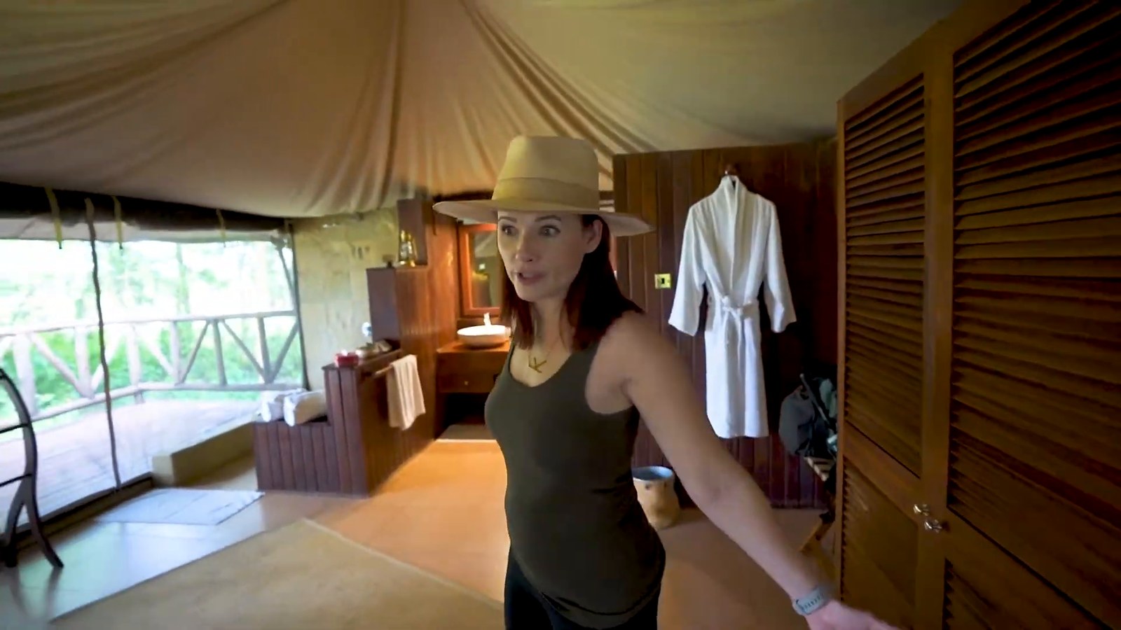 A woman in a hat stands in a tented bathroom with a vanity and basin, a white robe on a hook and open screens looking out to trees.