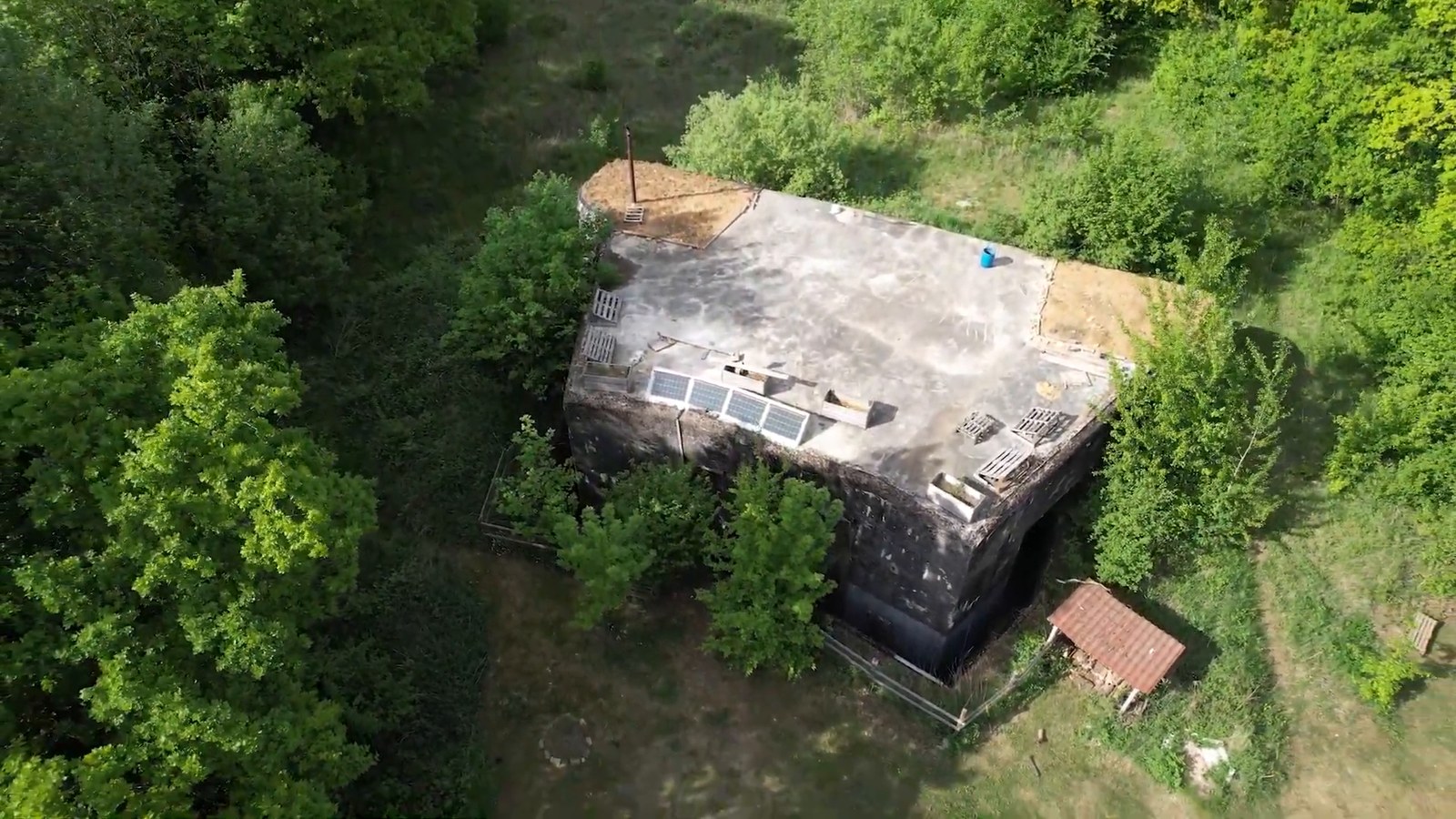 An aerial view of a concrete bunker in the woods with solar panels on its roof and a small wooden shelter beside it.