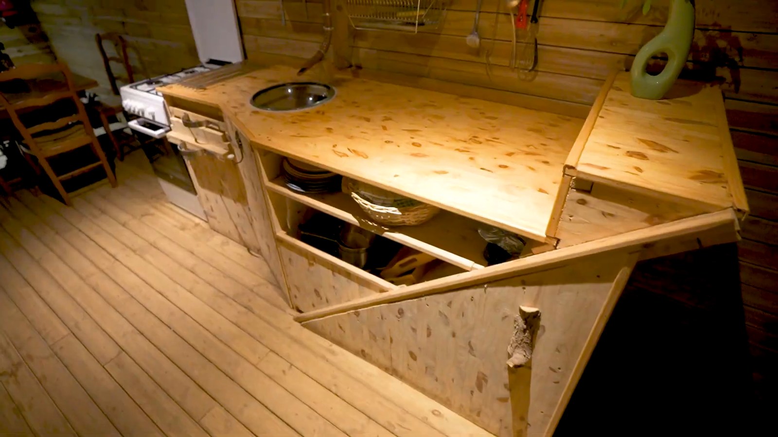 A pine kitchen counter with a round sink, a small stove and a dish shelf in a wood-paneled room.