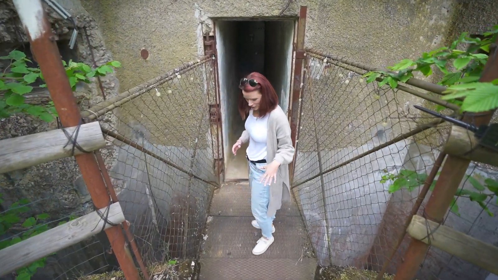 A woman in a grey cardigan steps out of a narrow concrete doorway onto a metal walkway between rusted fences at a bunker entrance.
