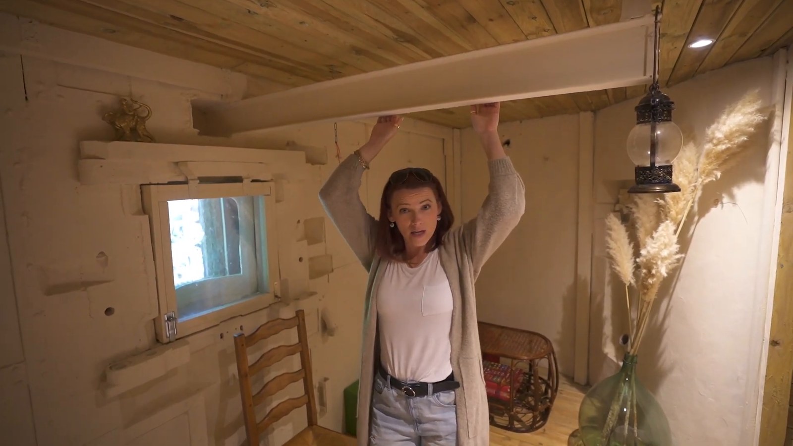 A woman reaches up to a steel beam running across the ceiling of a whitewashed room with a small window, a lantern and dried grasses.