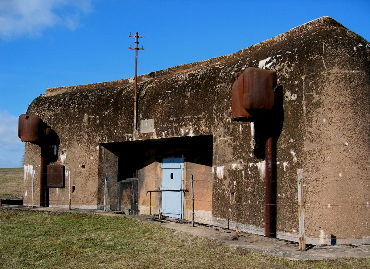 A weathered concrete Maginot Line casemate with two armoured cloches and a pale blue steel door under a blue sky.