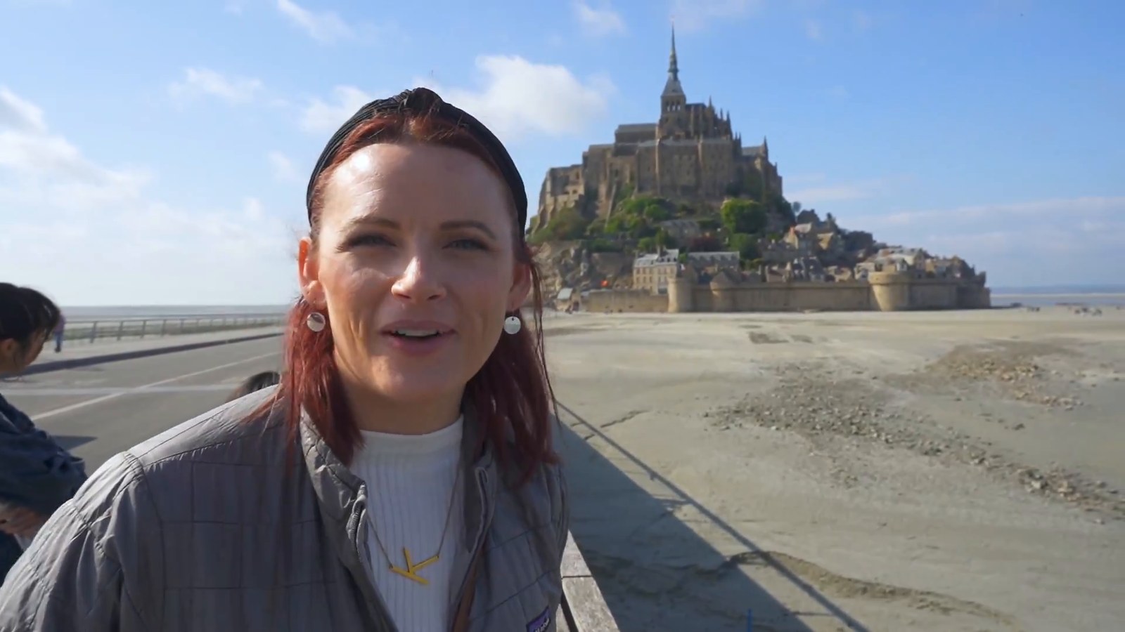 A woman smiles at the camera on the causeway with Mont-Saint-Michel rising behind her over the low-tide sands.