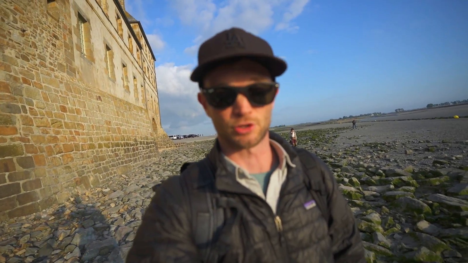 Kevin stands beside the old stone ramparts of Mont-Saint-Michel in low evening sun, with the wet bay stretching out behind him.
