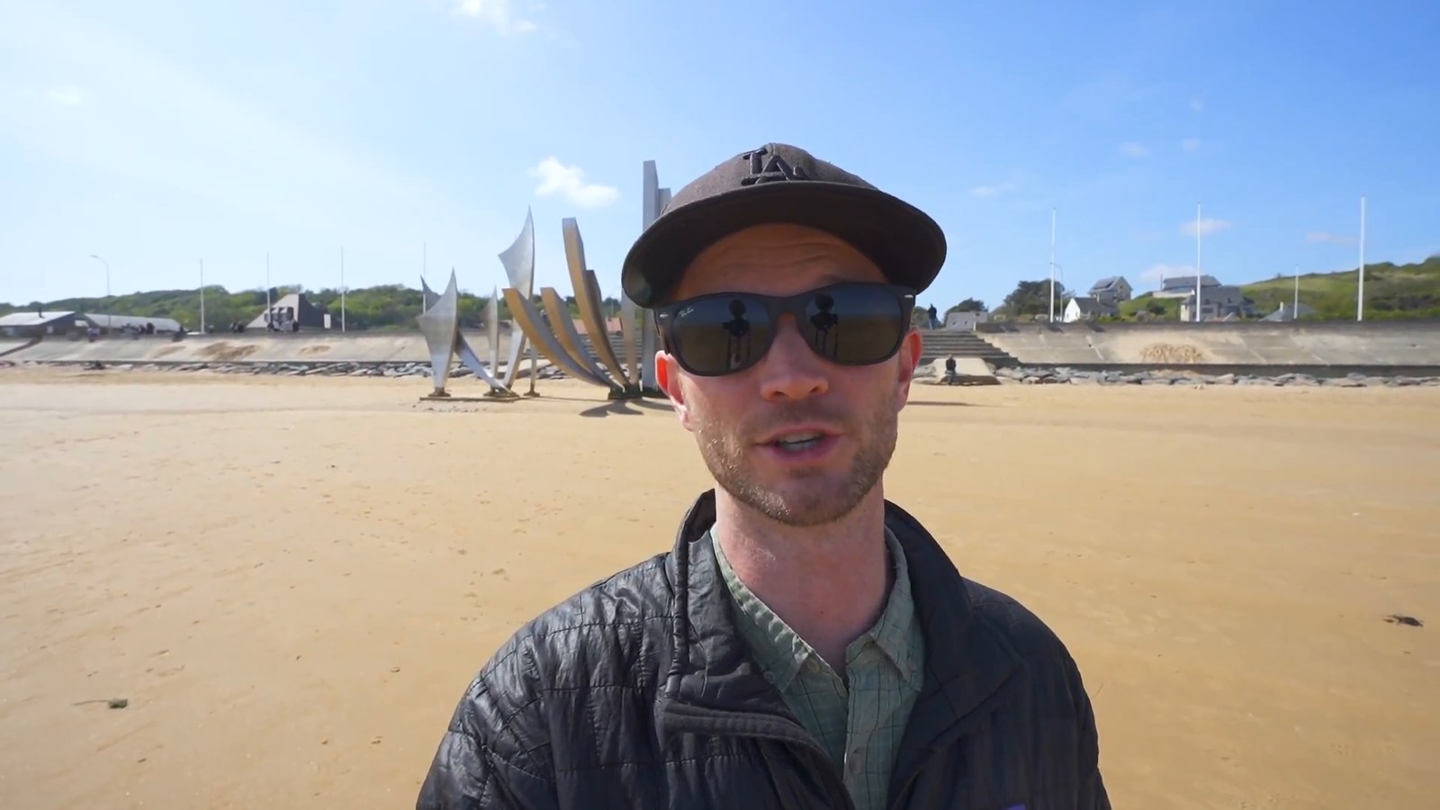 Kevin stands on the wide sandy expanse of Omaha Beach with the steel memorial sculpture behind him under a blue sky.