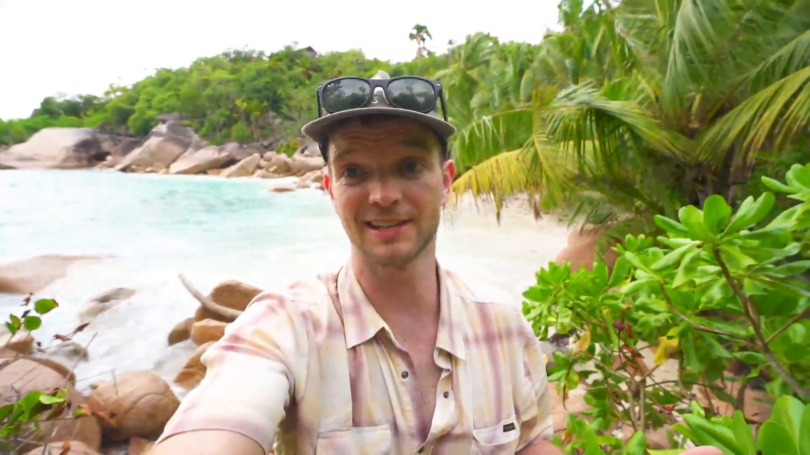 A man in a cap and sunglasses on his head takes a selfie in front of a small turquoise cove with granite boulders and palms.