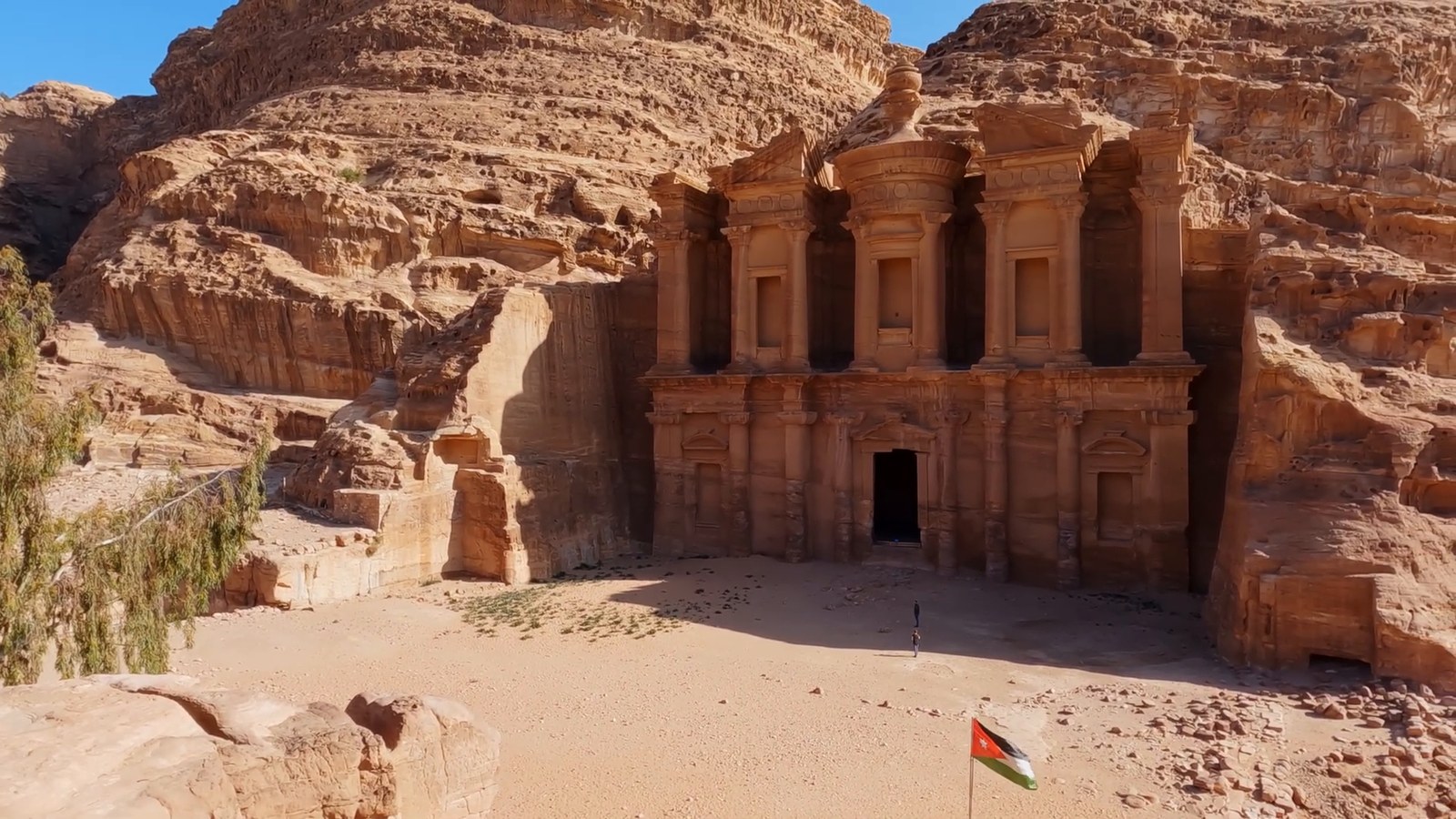 The Monastery at Petra seen from a rock ledge above, with its carved columns and doorway, an empty sandy square below and a small Jordanian flag.