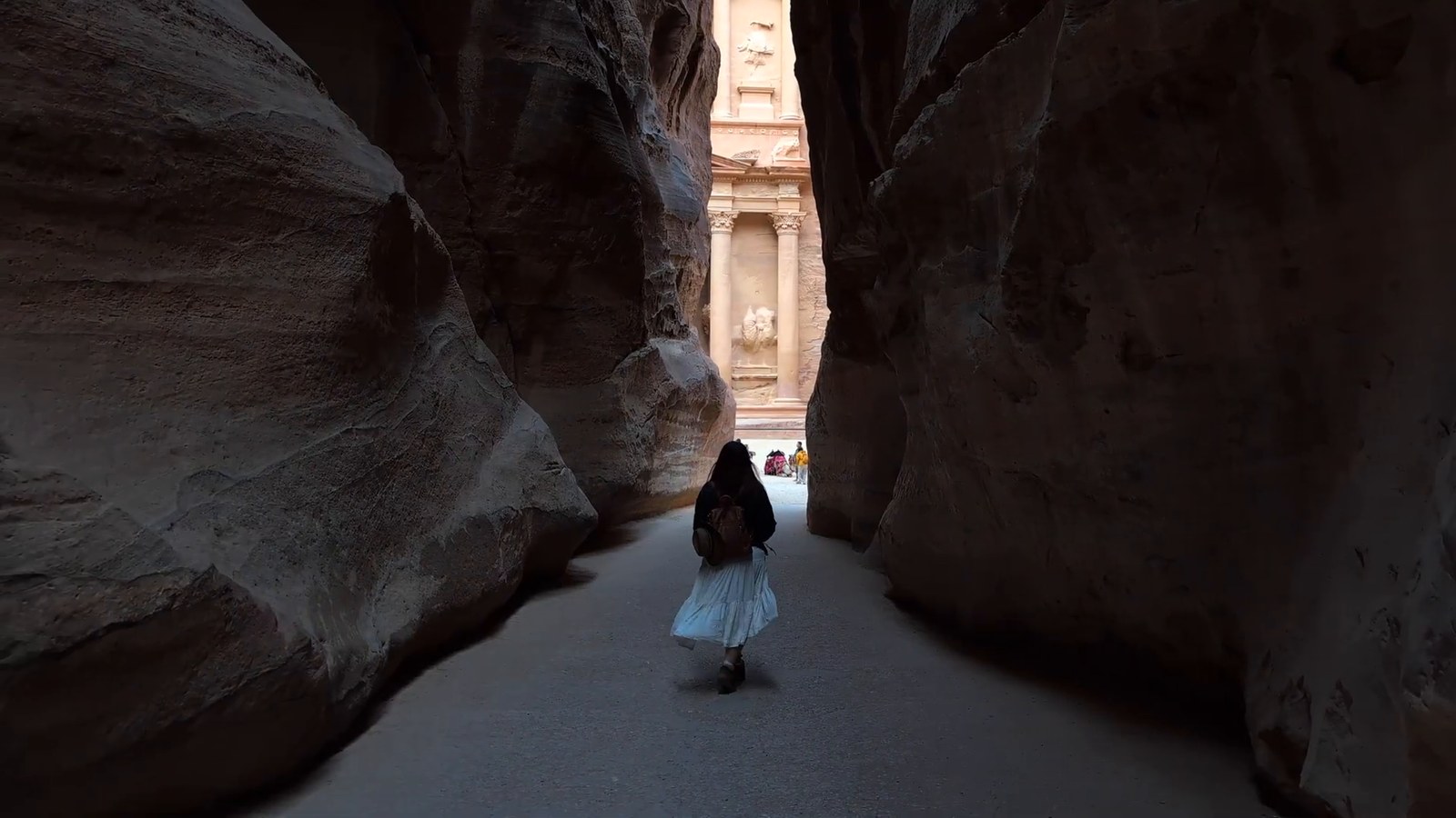 A visitor walks along the narrow Siq canyon toward the Treasury, glimpsed at the far end between two dark rock walls.