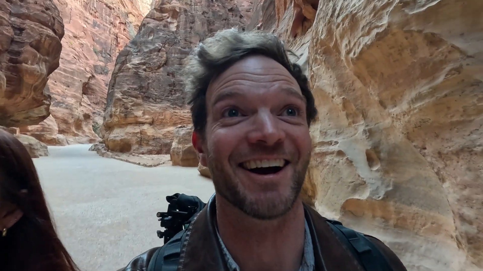 A man grins widely in the sandy Siq canyon, with pink-orange rock walls behind him.