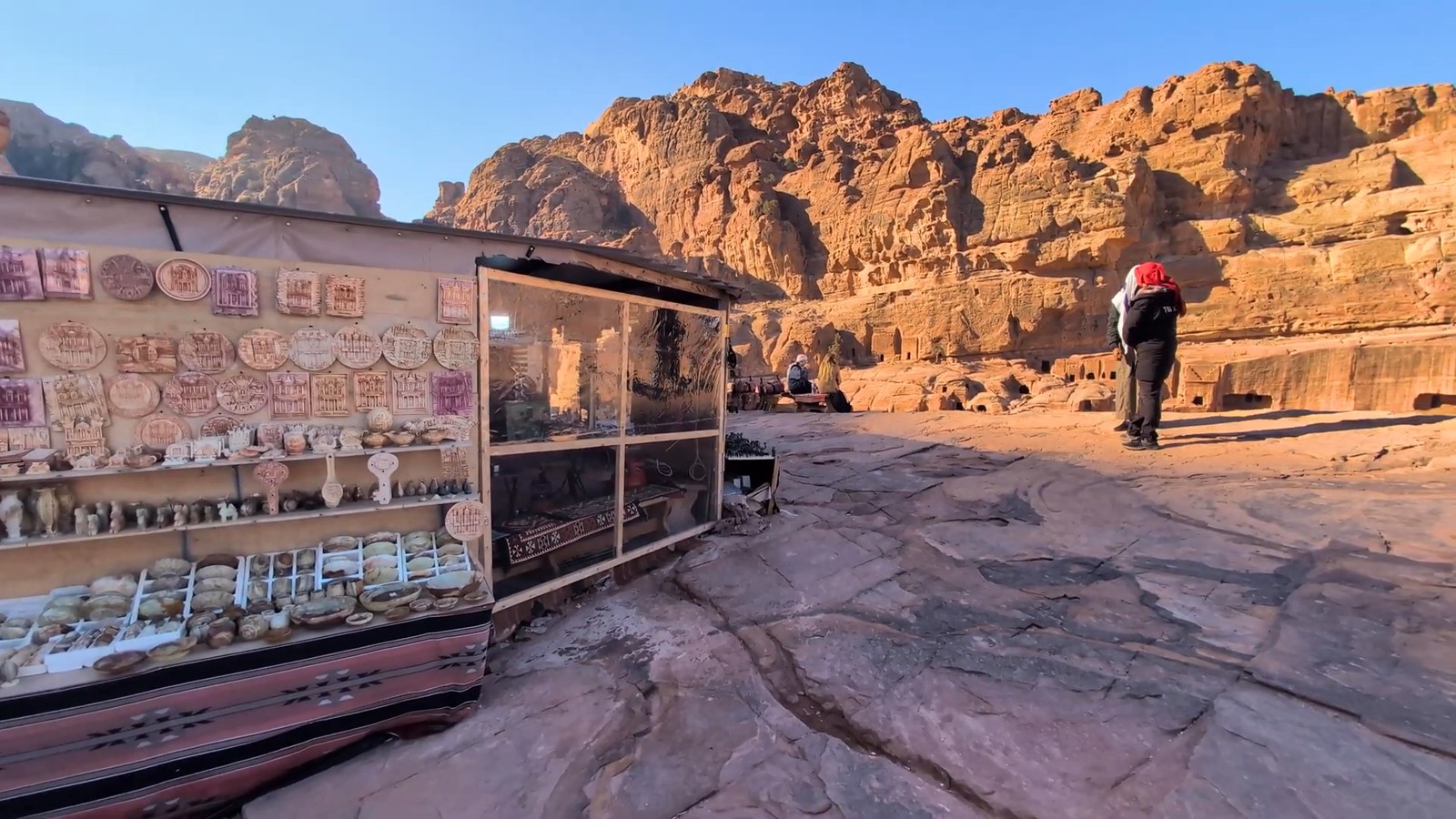 A small glass-fronted stall of souvenirs sits on a rocky ledge with red cliffs and tombs behind it under a blue sky.