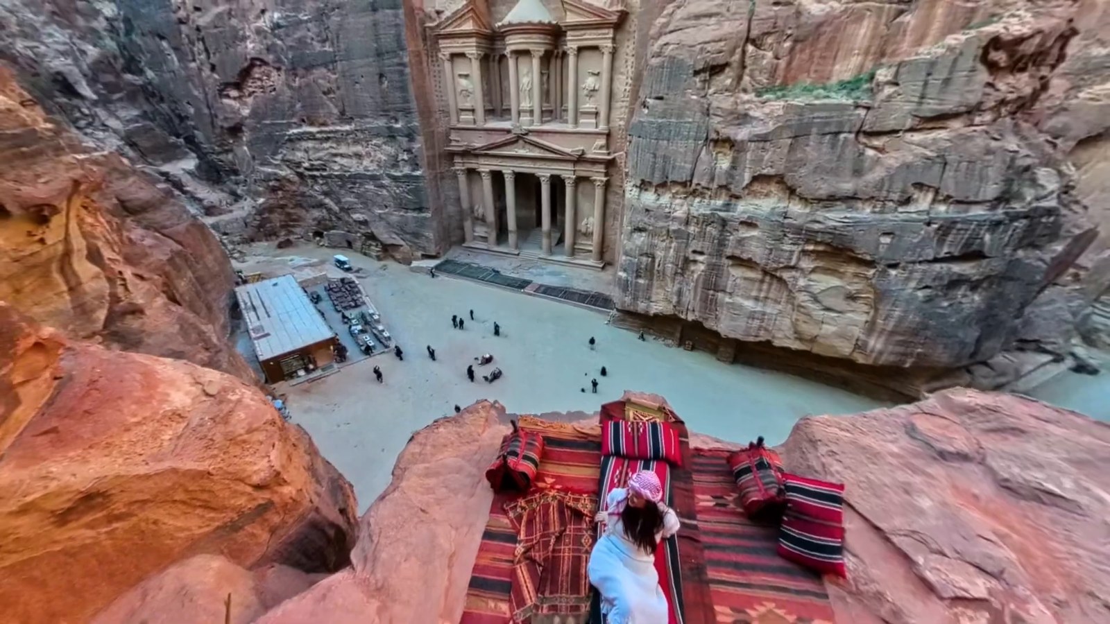 Looking down from a rug-covered rock ledge over the Treasury at Petra, with the sandy square and a few tiny visitors below.