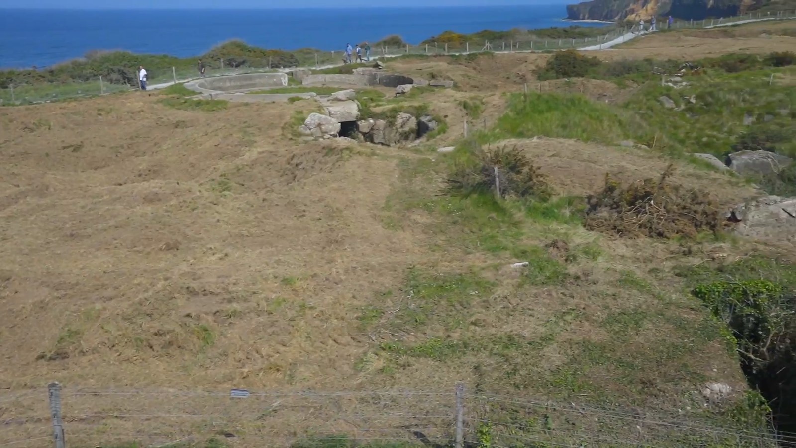 A grassy clifftop at Pointe du Hoc with uneven, cratered ground, a fence and the sea beyond.