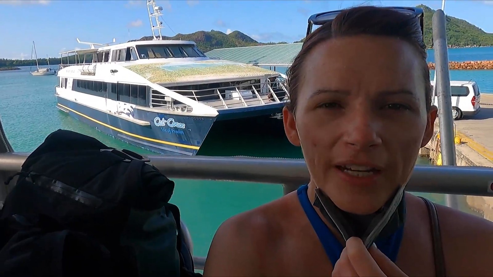 A woman with a backpack stands on the jetty in front of a white and navy passenger catamaran ferry with green hills behind.