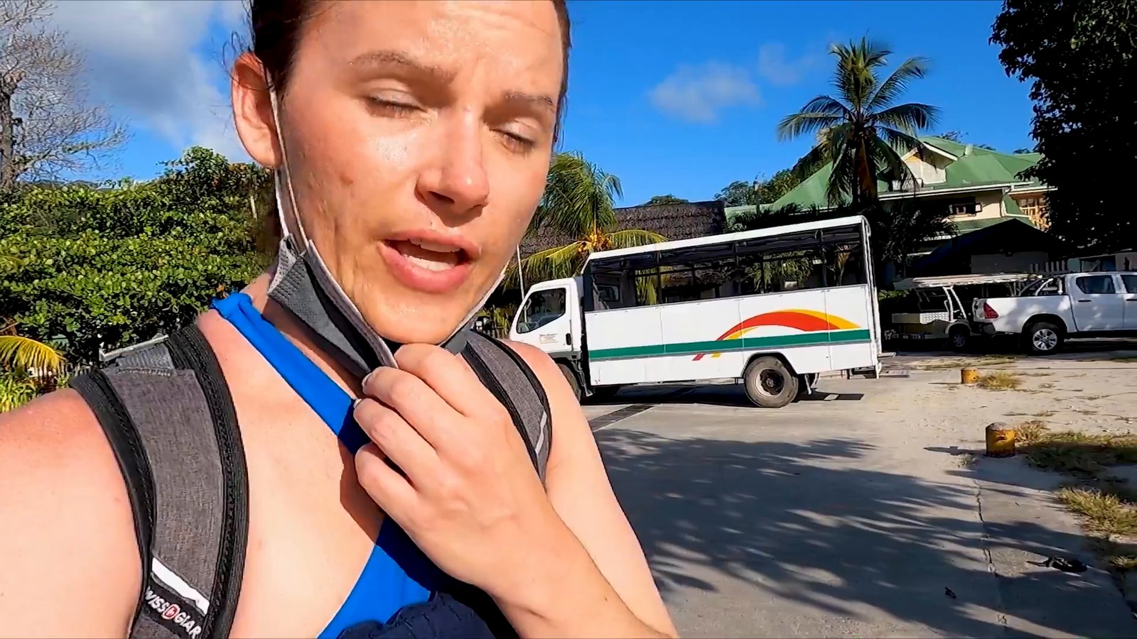 A sweaty woman wearing a face mask pulled down stands near a white public bus parked by palm trees.