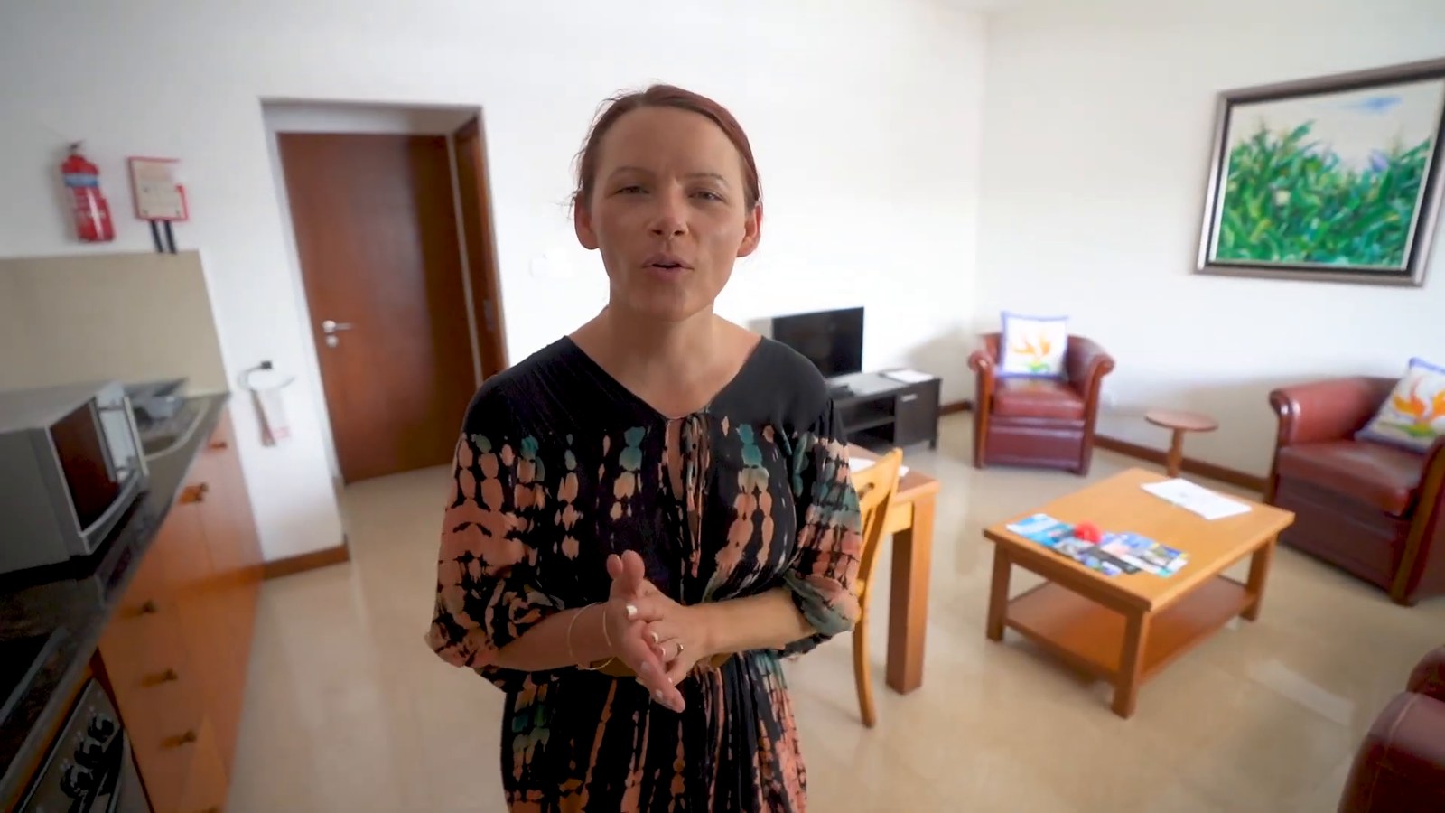A bright apartment living room with a leather sofa, wooden table and a small kitchen area, with a woman speaking to the camera.