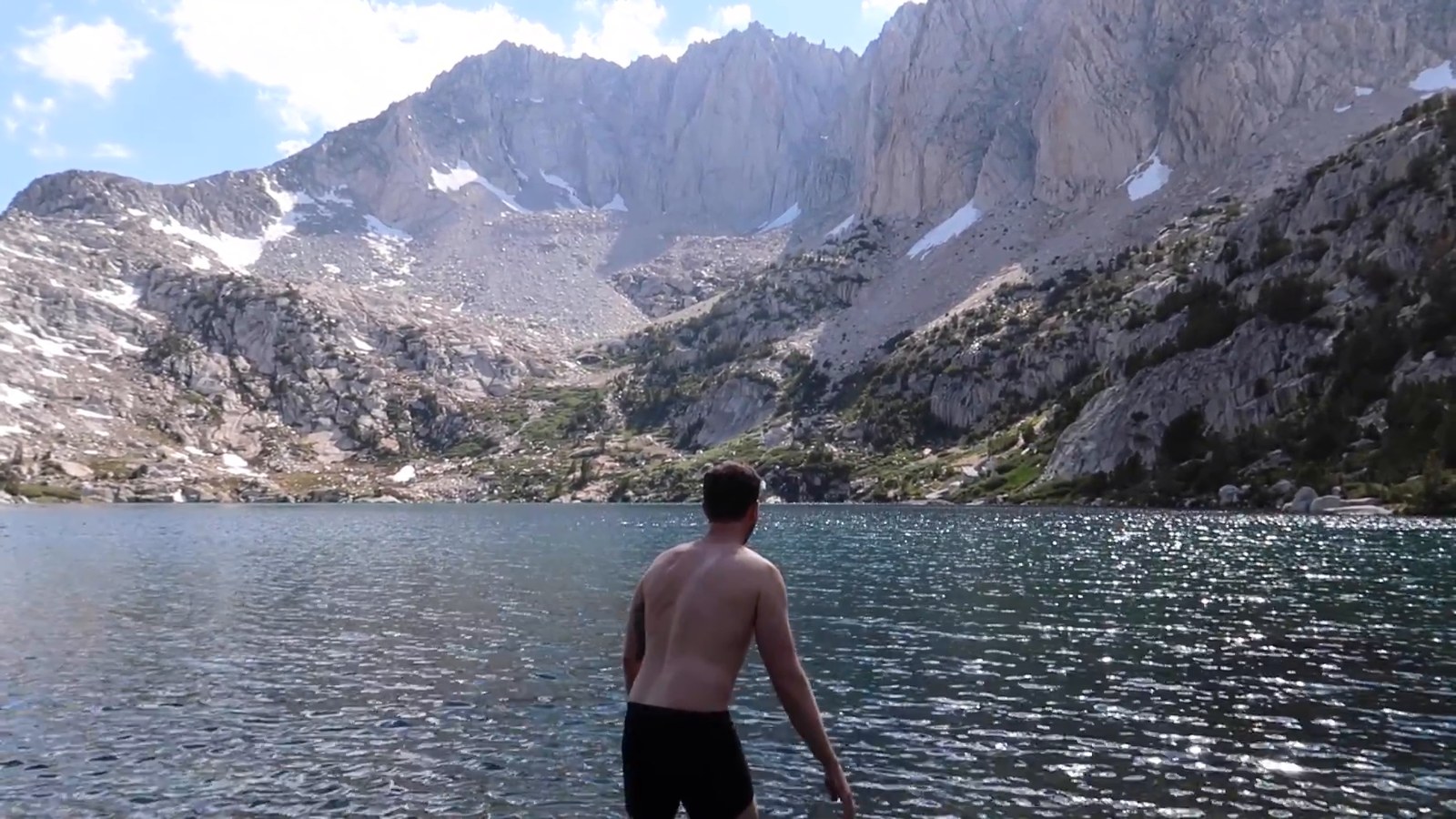 A person wades into a cold mountain lake with a rugged granite ridge rising behind it.