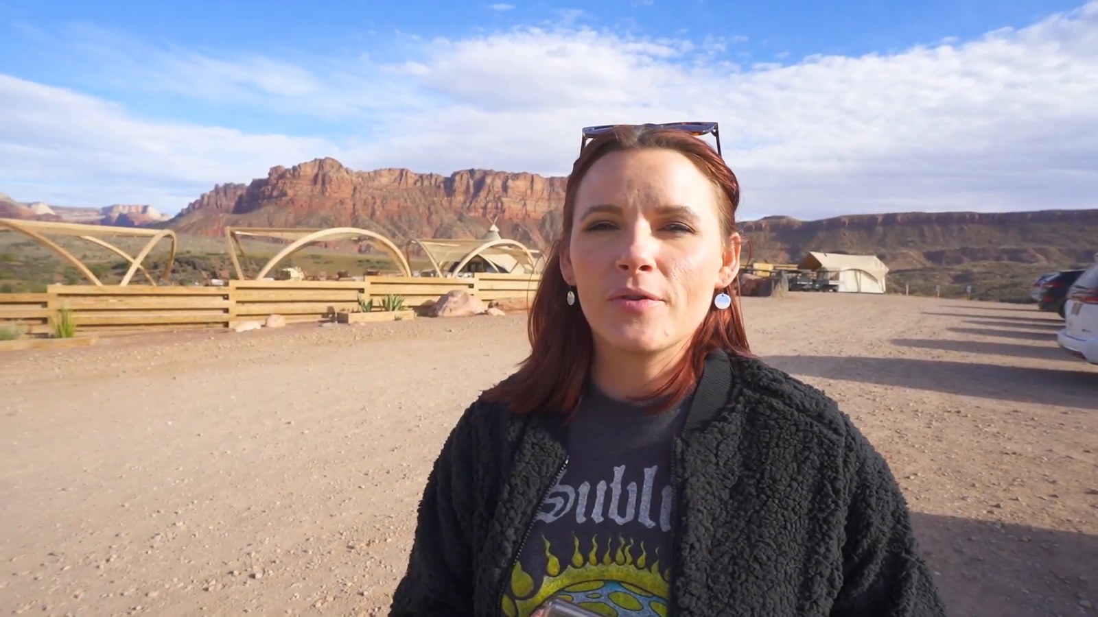 A woman with sunglasses on her head standing on a gravel lot, with red rock cliffs and wooden camp structures behind her.