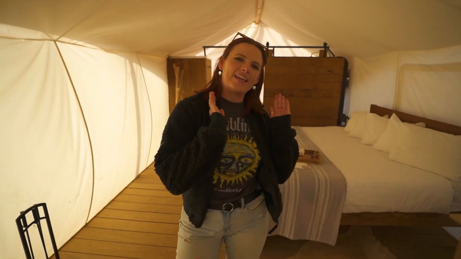 Inside a deluxe tent: a king bed with a striped throw, a wooden headboard and canvas walls, with a woman standing beside it.