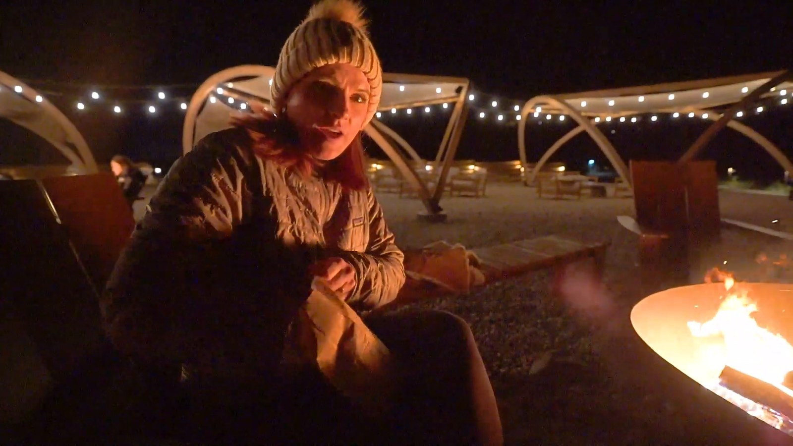 A woman in a woolly hat and puffer jacket beside a fire pit at night, with string lights in the background.