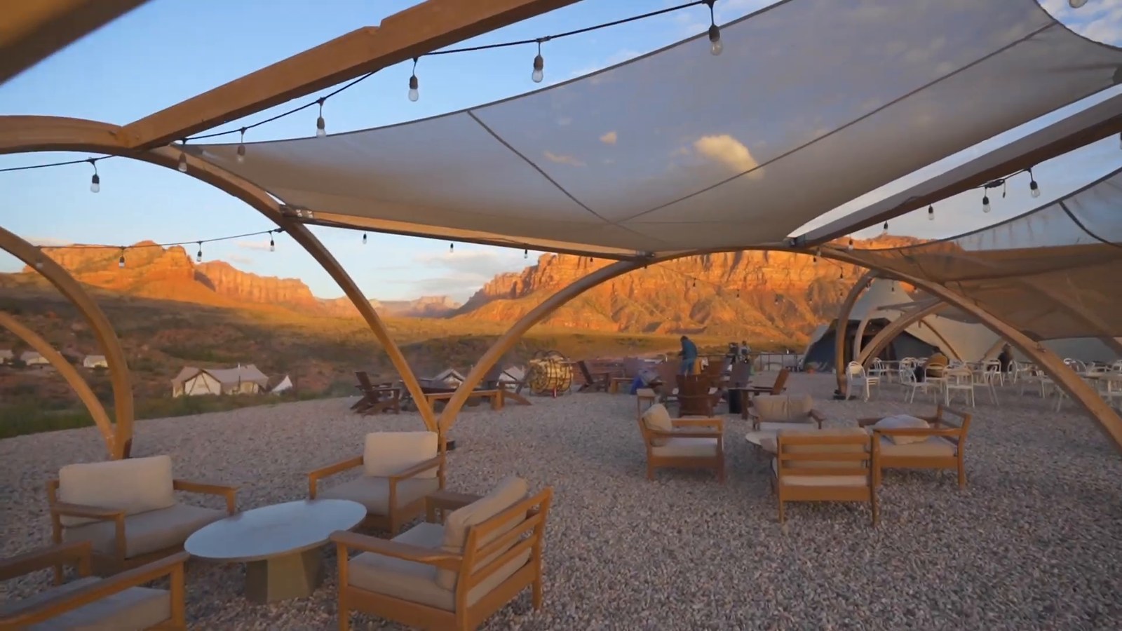 An outdoor lounge area under curved timber canopies strung with lights, with armchairs, a gravel floor and red cliffs lit by evening sun.