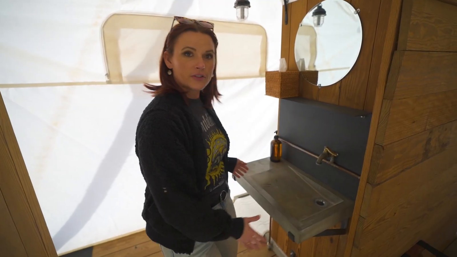 A woman beside a concrete sink with a brass tap, a round mirror and a wood-clad wall inside the tent bathroom.
