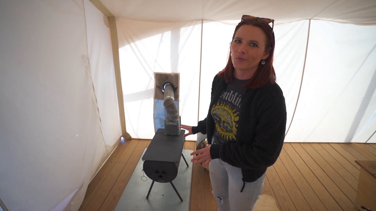 A woman beside a black wood-burning stove with a flue pipe inside the canvas tent.